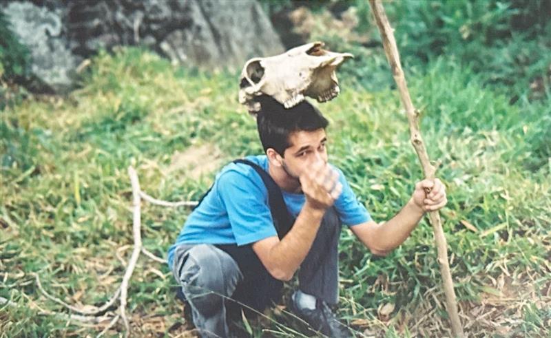 A imagem mostra Diogo Bercito, um homem branco, sentado na grama, segurando o pr&oacute;prio nariz. Ele veste uma camiseta azul e uma cal&ccedil;a jeans, e se apoia em um longo galho. Acima de sua cabe&ccedil;a, h&aacute; um cr&acirc;nio de um animal.